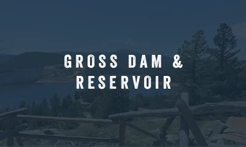 Gross Reservoir and dam surrounded by pine-covered mountains, viewed from an overlook with a wooden railing
