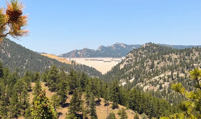 Gross Dam rising between rugged, pine-covered mountains beneath a clear blue sky