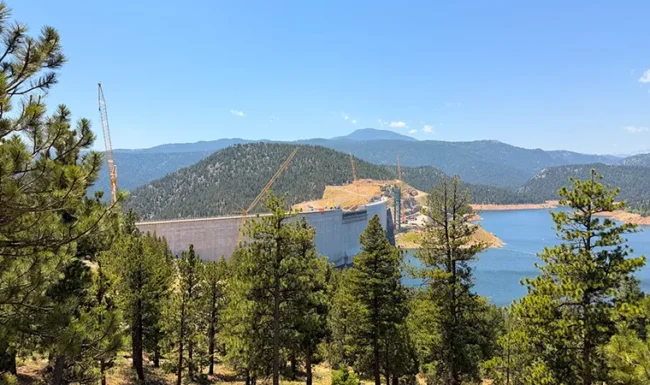 Gross Dam and construction cranes beside the blue waters of Gross Reservoir, framed by pine trees