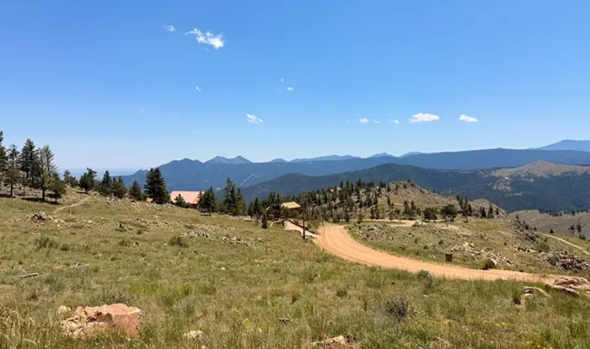 Grassy meadow, scattered pines and mountain homes beside Sunshine Canyon Drive, with distant ridgelines near Gold Hill, Colorado.