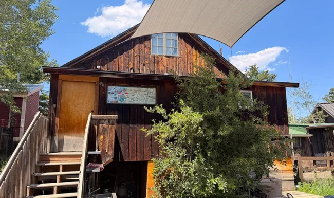 Rear exterior of Gold Hill General Store with wooden stairs, leafy shrubs and an overhead shade sail.