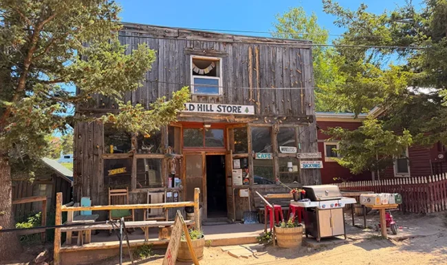 Weathered wooden storefront of Gold Hill General Store with an open entrance, cafe signs and outdoor seating.