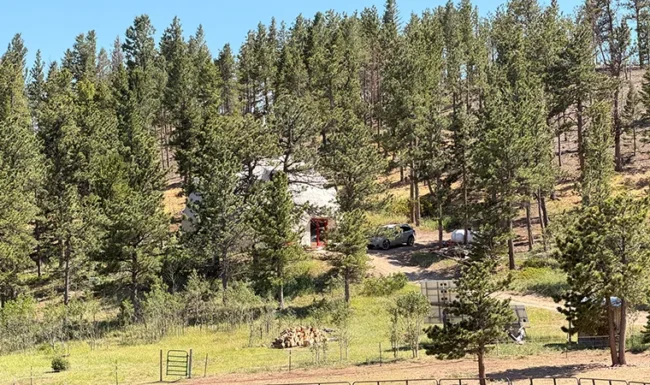 Geodome home nestled among pine trees on a hillside near Gold Hill, Colorado, with a fenced meadow in the foreground.